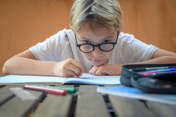 Cute blond child with glasses sitting at the table doing homework for school. young student intent on studying. boy concentrated in the study. interrogation preparation