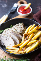 A skillet with roast pork meat served with potato fries and herbs on brown stone table. 