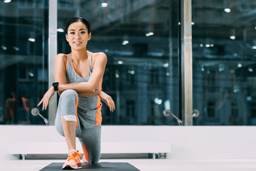 beautiful asian sportswoman smiling and doing stretch exercise on fitness mat in sports center