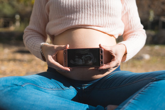 Pregnant woman showing a ultrasound image in her mobile phone. Concept of pregnancy, health care, motherhood, technology. Young mother waiting of the baby.