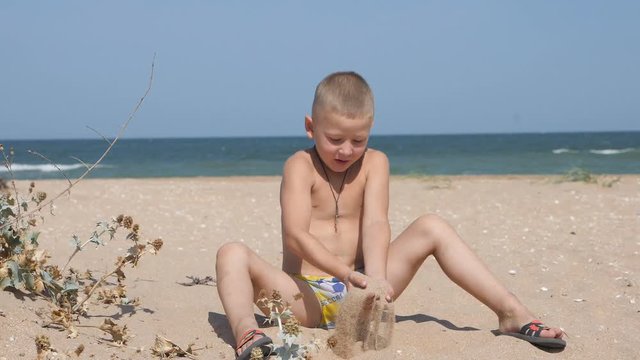 Blond boy sitting on the sandy beach throws up over his head the yellow sand.