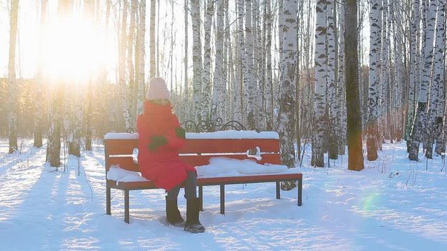 Pretty young woman in red warm jacket and felt boots sits on bench in pictorial city park with birches in beautiful winter frosty sunny day