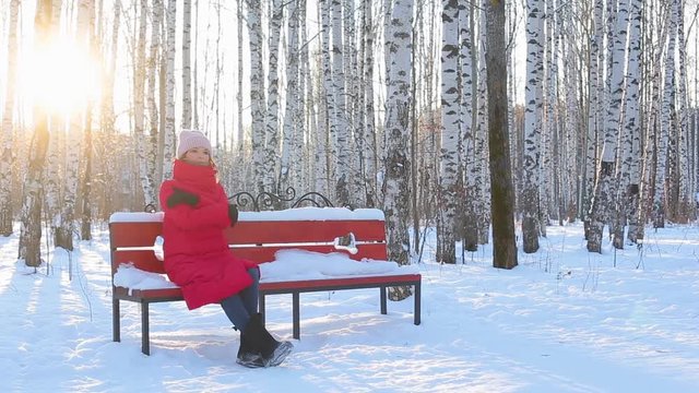 Pretty young woman in red warm jacket and felt boots sits on bench in pictorial city park with birches in beautiful winter frosty sunny day