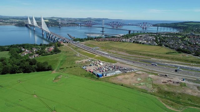 Aerial footage of traffic on the M9 motorway approaching and driving over the Queensferry Crossing bridge on the Firth of Forth with the old Forth Road Bridge and Railway Bridge beyond.