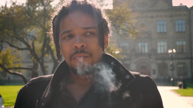 Close-up portrait of African-American guy with dreadlocks smoking on autumnal campus background.