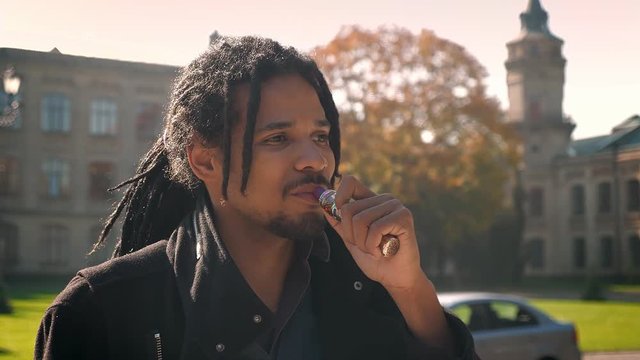 Closeup portrait of African-American guy with dreadlocks smoking and watching into camera on autumnal campus background.