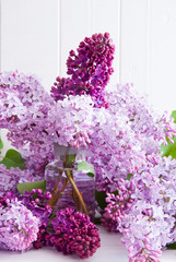 Purple flowers of lilac with leaves in a glass bottle on a white wooden background