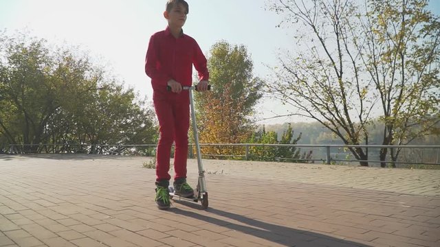 Cute boy in red shirt and pants riding a scooter in the park. The child actively spends time outdoors. Slow motion.