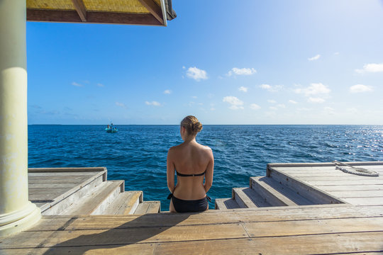 Junge Frau im Bikin sitzt an einer Treppe auf den Malediven und blickt ins Meer
