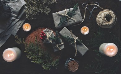 Yule winter solstice (Christmas) themed flat lay of gifts presents wrapped in gray paper vintage packaging with yarn. Dried flowers, evergreen tree branches, lit burning candles on dark wooden table