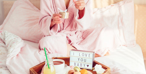 Breakfast in bed with I love you text on lighted box, coffee, macaroons on wooden tray and cropped woman in a bathrobe opening gift box. Birthday, Valentine's day morning. Wide banner. Copy space.