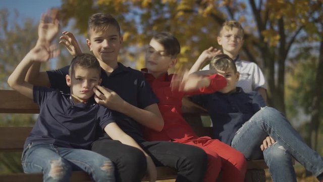 Five boys sitting on the bench and wave their hands on the camera. Friends spend time in big company outdoors.