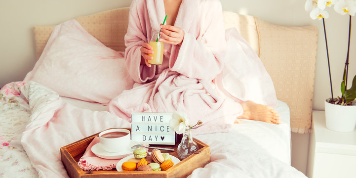 Good morning concept. Breakfast in bed with Have a nice day text on lighted box, coffee and macaroons on tray and blurred woman in bathrobe drinking juice. Hospitality, care, service. Copy space.
