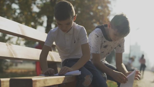 Two boys making paper planes outdoors. Kids sitting on the bench in the park