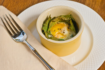 Top view, close distance of a yellow ramekin cup of a roasted egg, with fresh ground pepper, in bed of roasted kale on round, white plate with forn on wood serving board-paleo diet