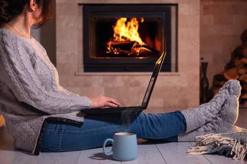 Young freelancer woman sits at the floor with a laptop on the fireplace background. 