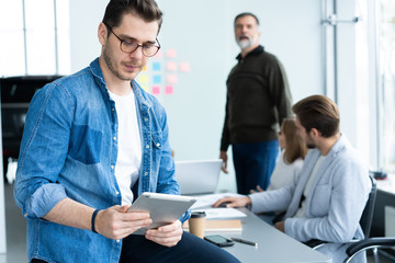 Young and creative. Handsome young man holding digital tablet and smiling while his colleagues discussing something in the background.