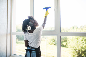Young african woman in yellow gloves cleaning window with spray detergent. Spring cleanup,...