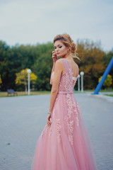 Fototapeta na wymiar Young beautiful woman in long pink evening dress walking path in park. Fashion style portrait of gorgeous beautiful girl outdoors