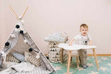Cute baby sitting at the table in the children's room in white, gray and blue tones. Near the...