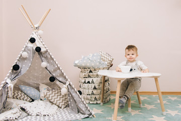 Cute baby sitting at the table in the children's room in white, gray and blue tones. Near the...