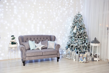 New Year's Decor in a photo studio in gray and white. Classic gray sofa with pillows, snow-white Christmas tree, gifts and a lantern, against the white wall with a garland