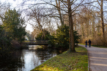 Outdoor sunny tranquil scenery landscape of couple tourist walk along walkway on waterside of frozen canal or lake in Park Sanssouci in winter season, and background of farther Chinese tea house.