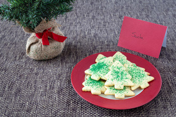 Christmas Tree shaped sugar cookies, with green sprinkles, on a red and white plate set out for Santa, and artificial tree on a brown background