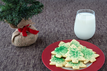 Christmas Tree shaped sugar cookies, with green sprinkles, on a red and white plate next to a glass of milk set out for Santa, and artificial tree on a brown background