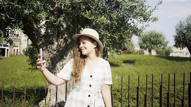 Young cheerful lady in dress and hat using smartphone in modern park in old city centre. Portugal, Porto