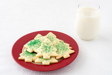 Christmas Tree shaped sugar cookies, with green sprinkles, on a red and white plate next to a glass of milk set out for Santa, on a white background