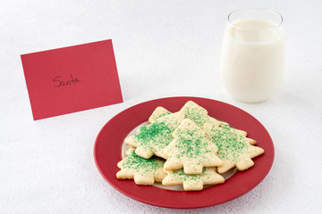 Christmas Tree shaped sugar cookies, with green sprinkles, on a red and white plate next to a glass of milk with a note set out for Santa, on a white background