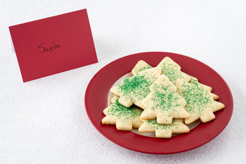 Christmas Tree shaped sugar cookies, with green sprinkles, on a red and white plate with a note set out for Santa, on a white background