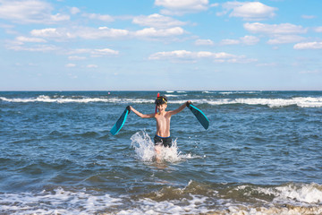 Happy teen boy in the swim flippers and snorkeling mask running оn the wave of the sea during summer vacation in the tropical resort town