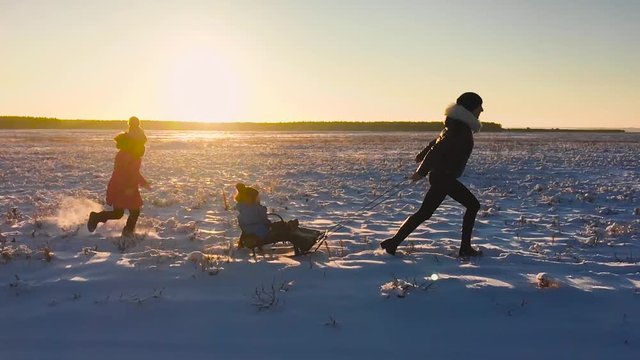 Teen daughter and mother pulling little son on sledge, running. Silhouette family playing in winter time. Active healthy lifestyle concept.