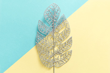 Silvery glittering monstera leaf on pastel blue and yellow background. Festive decoration. Tropical Christmas concept. Flat lay, copy space.