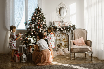 Young mother with two little daughters decorate a New Year's tree in the light cozy room with armchair and fireplace
