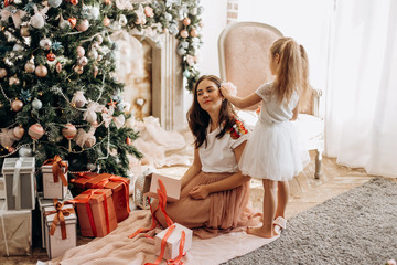 Happy young mother and her  little daughter in nice dress sit near the New Year's tree and open  New Year's gifts in the light cozy room