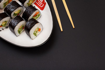 Sushi rolls in nori seaweed sheets on plate with wooden sticks on black background.