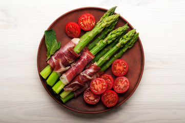 Asparagus with ham, tomatoes and egg poached in a in a clay plate. White wooden background.  Light diet breakfast.