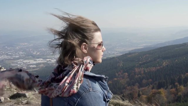 Young woman wearing sunglasses in bright sunlight in strong mountain wind blowing her hair overlooking landscape