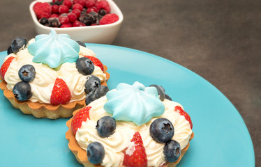 Two tartlets with white butter cream, blue marshmallow on top and strawberries and blueberries on blue plate and cup with raspberries and currants near the plate. Side view