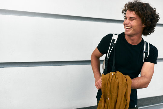 Outdoor image of handsome smiling man with curly hair with backpack standing at building wall on the street while waiting his colleagues. Businessman wears casual black t-shirt. Copy space for text