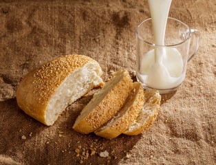 A bun with sesame seeds and a glass of milk on the burlap.  Milk pours into the glass.