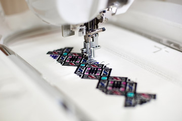 Close-up of sewing machine creating colorful abstract geometrical pattern on white fabric on blurred background. Modern technology, fashion and traditional clothing manufacturing concept.