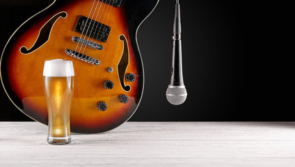 Glass of beer and microphone near electric jazz guitar on white wooden desk. Dark background