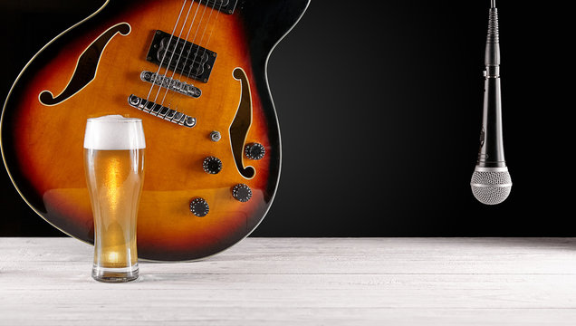 Glass of beer and microphone near electric jazz guitar on white wooden desk. Dark background