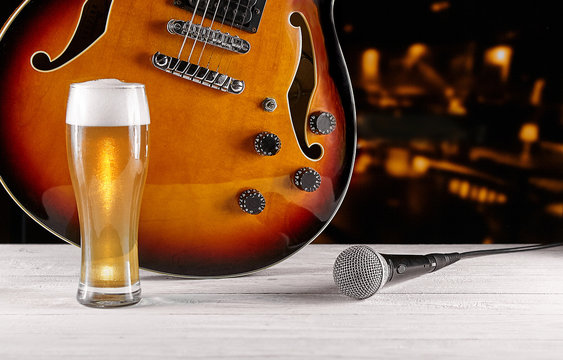 Glass of beer and microphone near electric jazz guitar on white wooden desk. Dark background