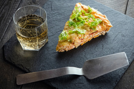 slice of pizza on slate plate with metal spatula and drink on wooden table.