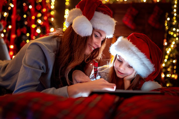 Fototapeta na wymiar Christmas time. Smiling family mother and daughter in santas hats and pajamas watching funny video or choosing gifts on digital tablet while lie on the bed
